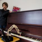 Marius Schmeding lehnt am Klavier und sieht dem Roboterarm beim Spielen zu. Foto: Holger Vogel